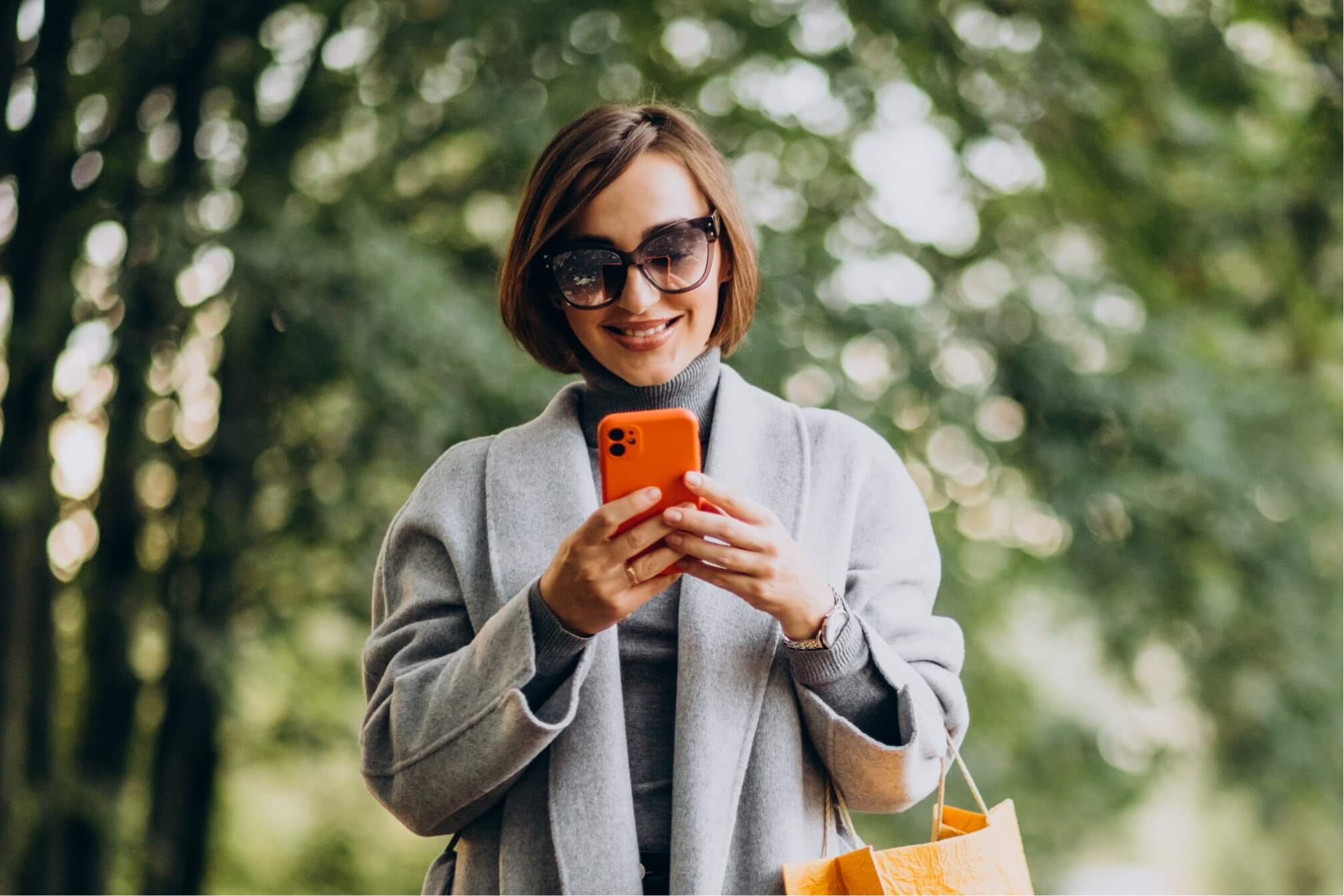 Młoda, uśmiechnięta kobieta w parku spogląda na wyświetlacz pomarańczowego smartfona.